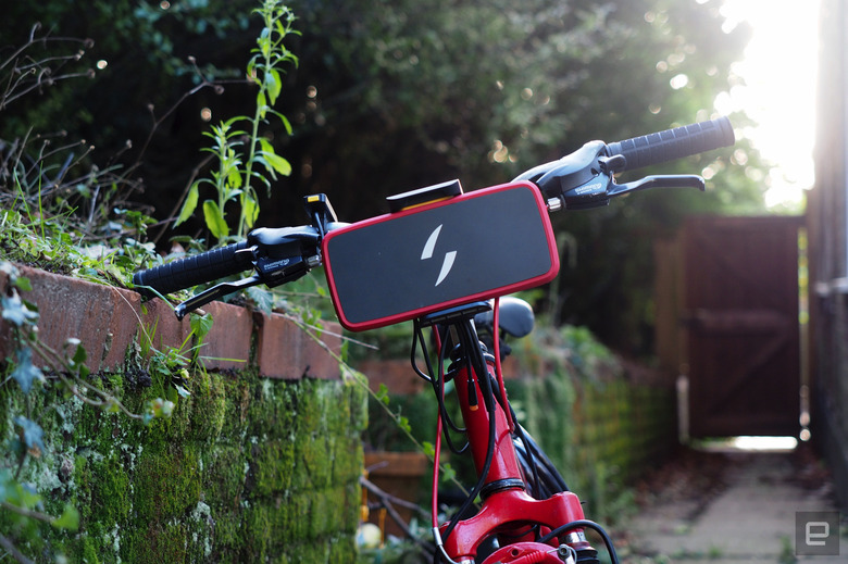 The front view of a red bike with the Swytch Air e-bike conversion kit installed, showing the battery pack portion attached to the front of the handlebars. The bike is leaning against a vine-covered wall.