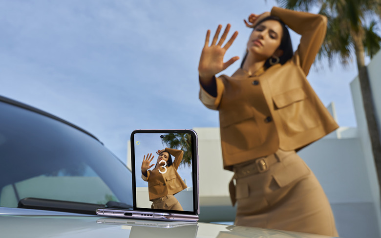 A half-opened Tecno Phantom V Flip 5G phone placed on a car hood, with its main camera facing a model who is posing with a hand gesture to remotely trigger the shot.