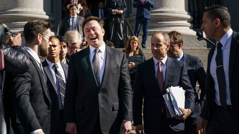 A suit-clad Elon Musk cackles on courthouse steps, surrounded by men in suits.