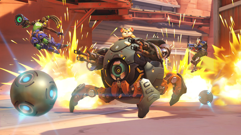 A still from Overwatch 2 showing the character Wrecking Ball with an explosion behind his vehicle and two other characters flying through the air