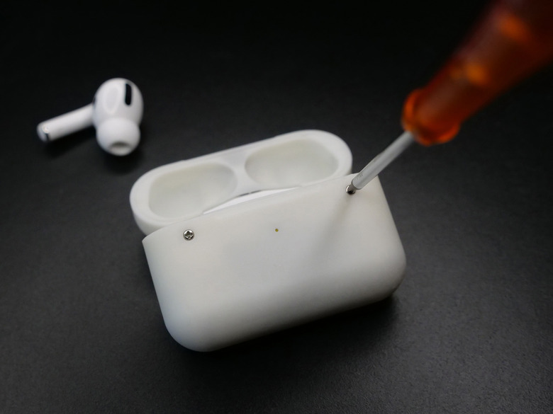 A custom AirPods Pro case with screws. A screwdriver is unscrewing one of the two visible screws. A lone AirPods Pro bud sits on the dark surface behind it.
