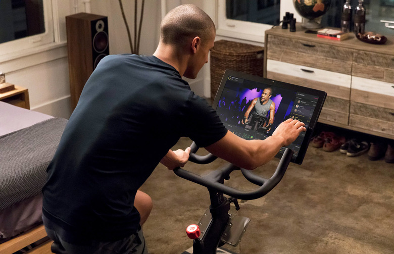A promotional image of a man in his room riding a Peloton bike with a touchscreen display in front.