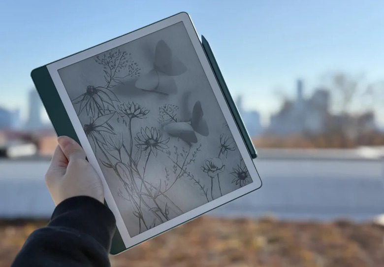 A Kindle Scribe with a sketch of flowers on it is pictured held up against a blurred outdoor background