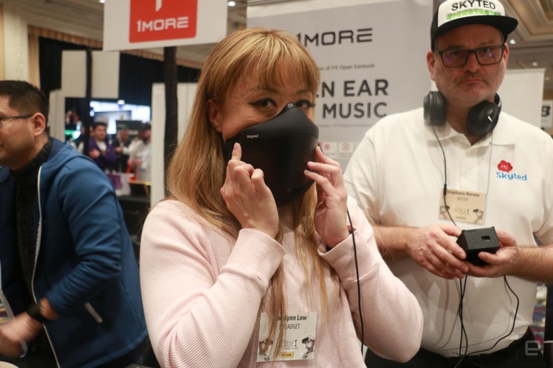 A person holding the Skyted mask to her face at a busy event with people and banners in the background.