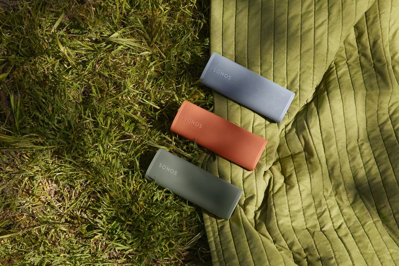 Three Sonos Roam 2 portable speakers (green, red, blue), sitting side-by-side (diagonal to the camera) on an outdoor blanket on top of grass.