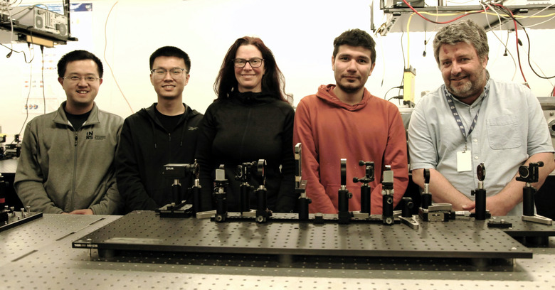 Group photo of five members of a research team focused on a scientific camera. They stand smiling in a laboratory with various imagine equipment on the research table in front of them.