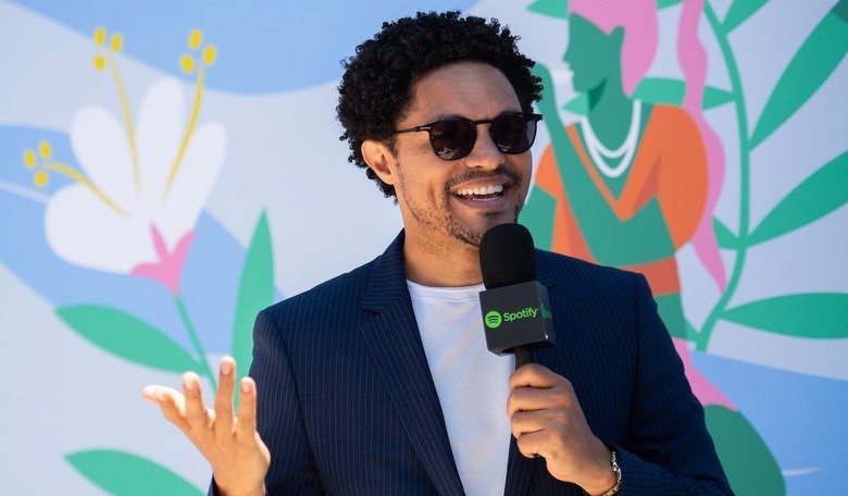 Trevor Noah holds a microphone with Spotify logo on it and a flowered background behind him. 