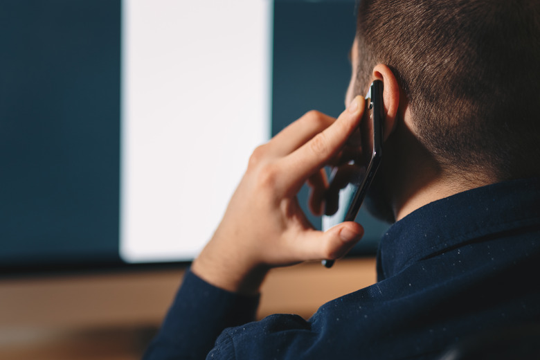young businessman working in his home office, using his mobile phone while sitting in a chair in front of his personal workspace, looking at the screen.