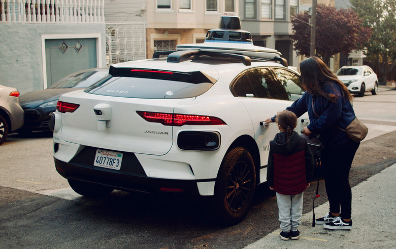 A woman and a child and are standing next to a white autonomous vehicle, in the process of pulling a door open. 