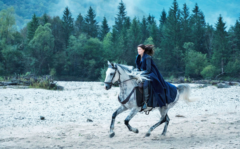 A still from Amazon's 'Wheel of Time' TV series, showing Rosamund Pike riding on a horse over sand and gravel with trees in the background.