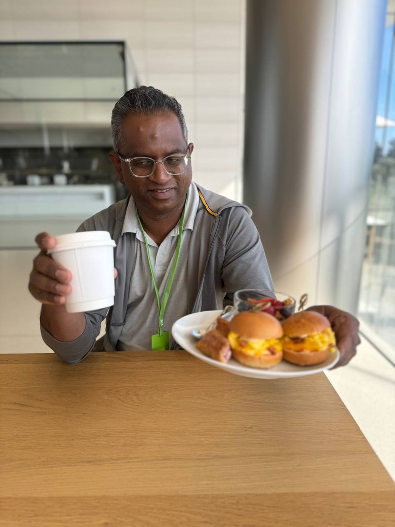 Senior editor Devindra Hardawar holding up a coffee cup in his right hand and a plate of breakfast foods in his left.