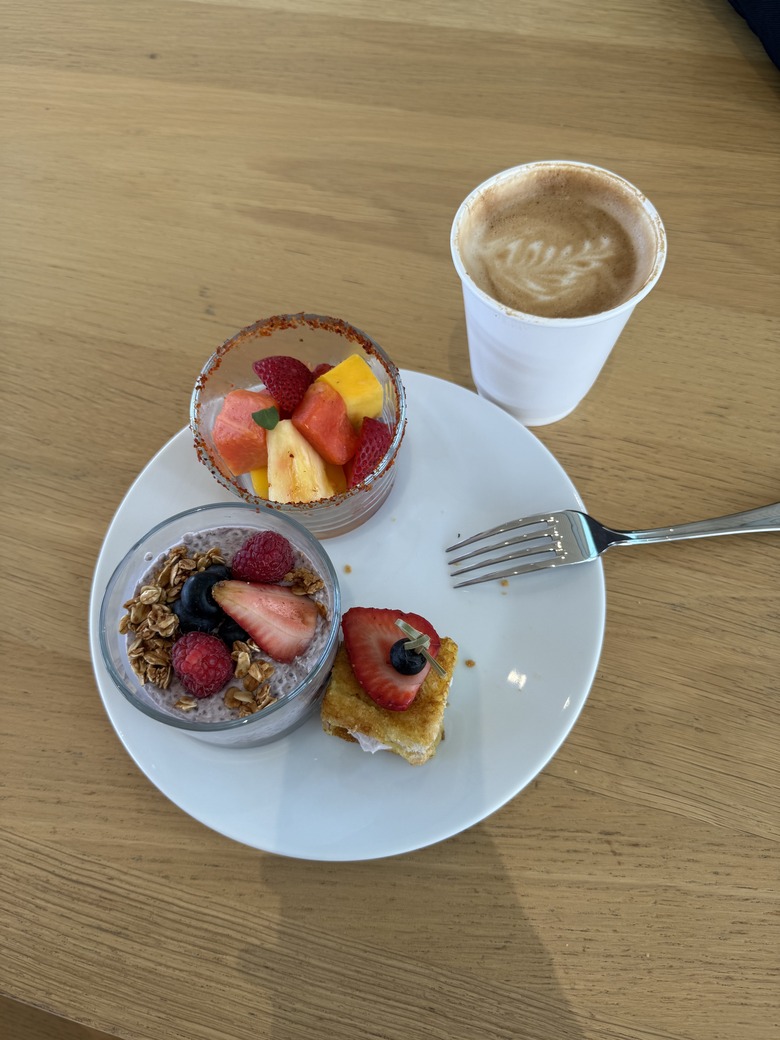 A plate of food with three mini items -- a cup of mixed fruit, a cup of granola and fruit parfait and a small french toast. A small cup of coffee sits next to the plate.