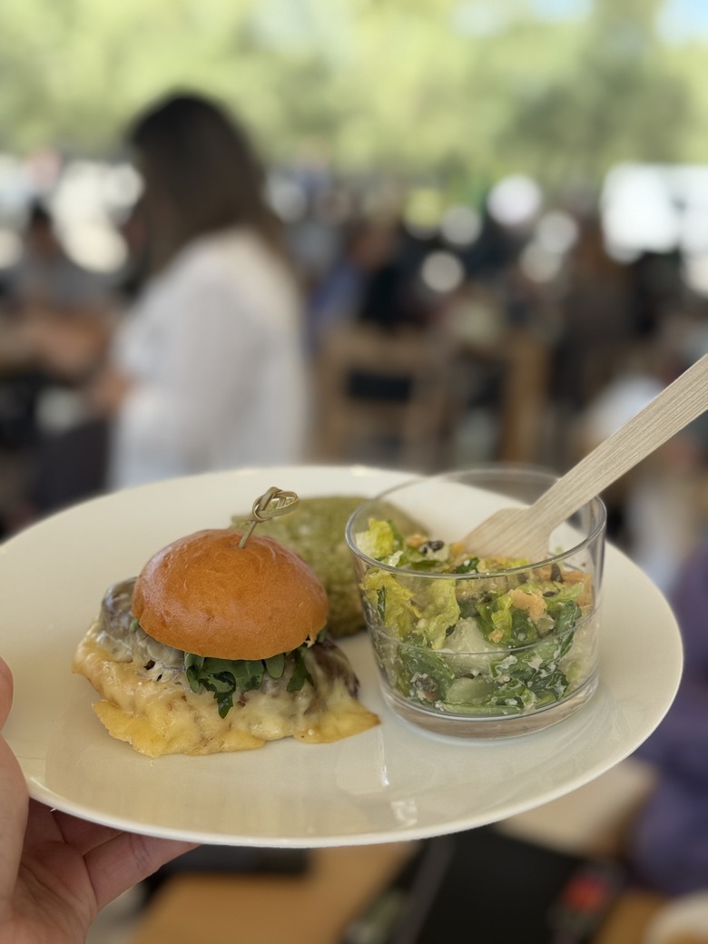 A plate held in mid-air. On it sit a beef slider, a transparent cup with salad inside and a green cookie.