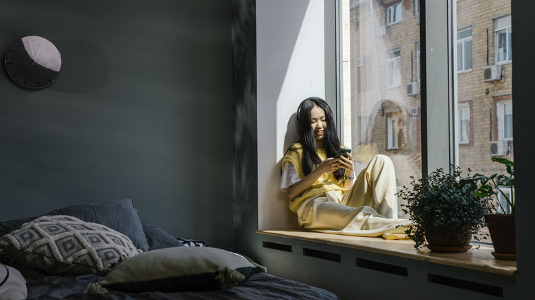Asian woman sitting by a window indoors, smiling at her phone with headphones on in a cozy home setting.