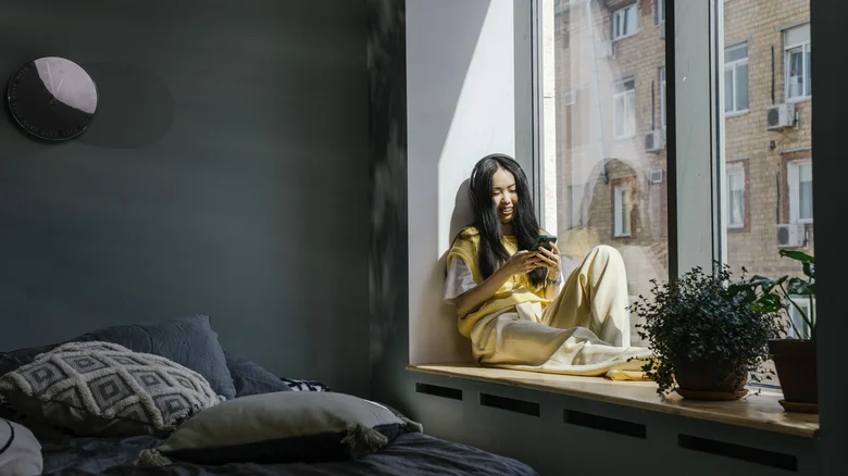 Asian woman sitting by a window indoors, smiling at her phone with headphones on in a cozy home setting.