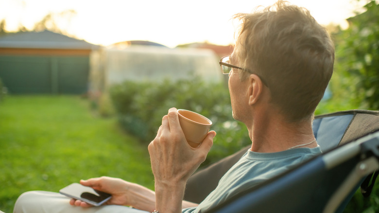 Man sitting in backyard with smart phone with a mug in his hands.