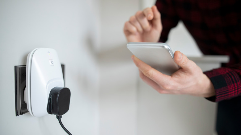 A person uses a smartphone beside a smart plug connected to a wall outlet.