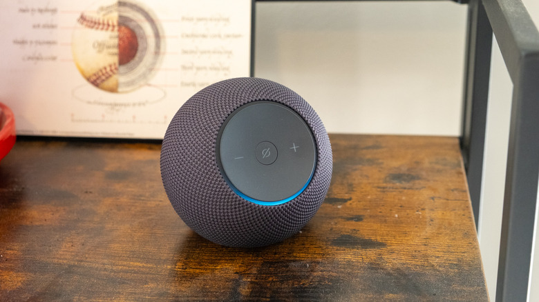 A black Amazon Echo Dot Max speaker sitting on a shelf with its blue light indicator glowing.
