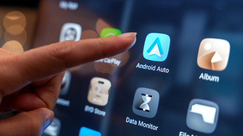 A close-up of the Android Auto app on a car infotainment screen, with a finger approaching to tap it.