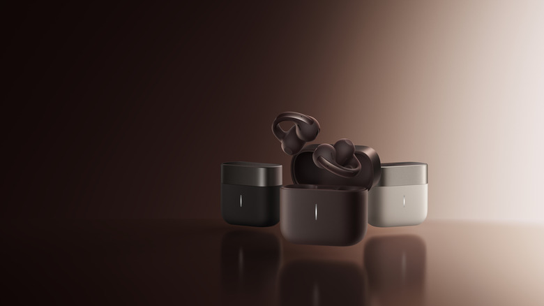 Photo of the Anker Soundcore AeroClip 2 Pro earbuds. A render of the brown earbuds above an open case in front of one black and one white case. Background is a dramatic, monochrome spotlight gradient.