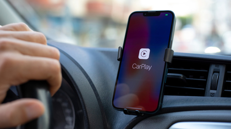 iPhone mounted on a car dashboard displaying the Apple CarPlay connection screen while a driver holds the steering wheel.