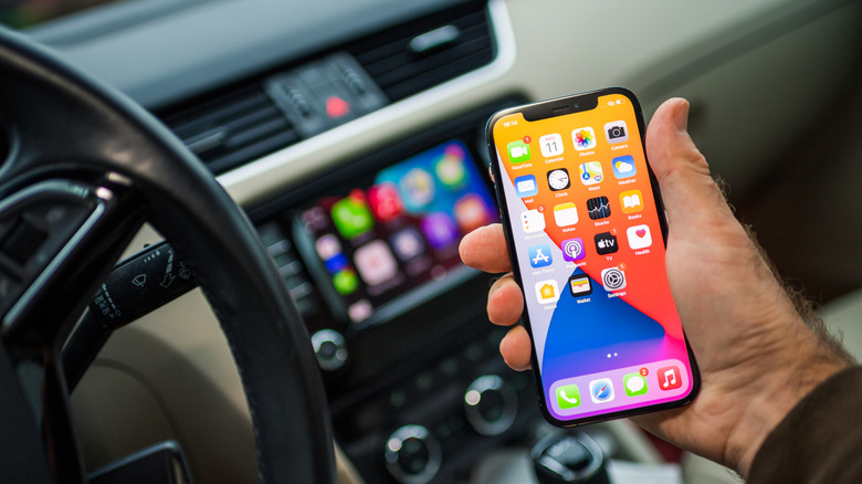 A hand holding an iPhone with a blurred CarPlay display in the background.