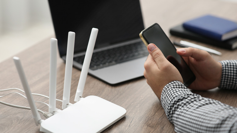 A man uses a smartphone next to a laptop and router.