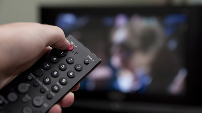A close-up view of a TV remote, with an blurred image of a TV towards the back.