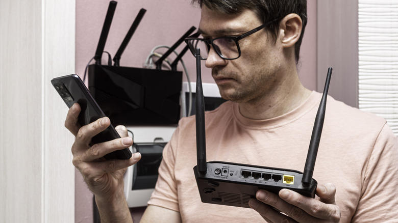 Man holding a Wi-Fi router in one hand and smartphone in another hand, in front of networking equipment.