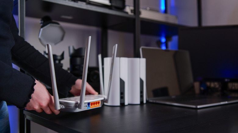 A person placing a white Wi-Fi router with external antennas on a dark desk next to two Wi-Fi routers and a laptop.