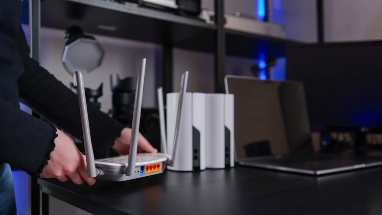 A person placing a white Wi-Fi router with external antennas on a dark desk next to two Wi-Fi routers and a laptop.