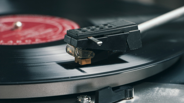 A cartridge and stylus play upon the grooves of a black vinyl record with a red label.