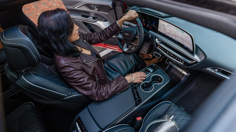 A woman in a leather jacket and skirt sits in a Cadillac Lyriq's sleek black leather interior, looking at car's dashboard where a city map is visible.