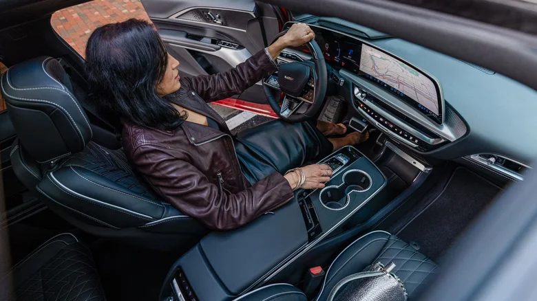 A woman in a leather jacket and skirt sits in a Cadillac Lyriq's sleek black leather interior, looking at car's dashboard where a city map is visible.