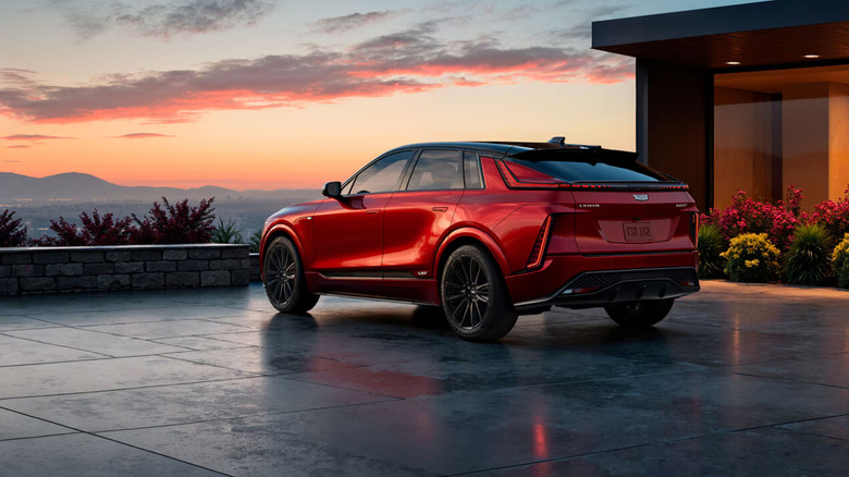 A red Cadillac Lyriq sits parked in the driveway of a modern home, with a red and purple sunset descending over distant mountains.