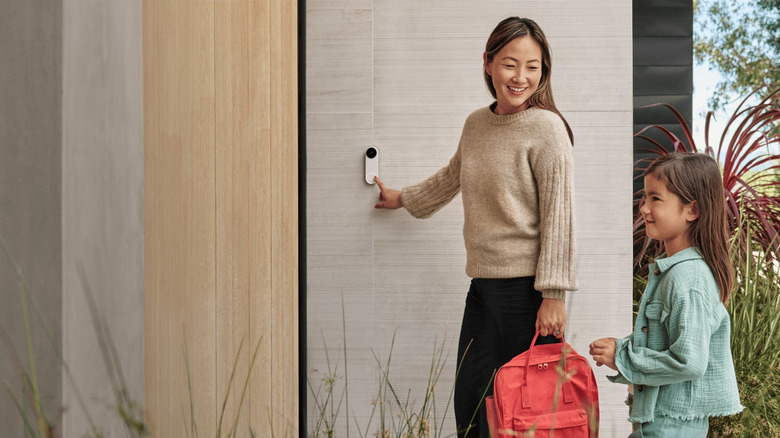 An adult presses the buzzer on a video doorbell device that that's attached to a wall while smiling at a child.