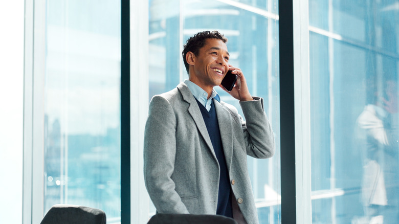 A man standing by the window in his office with a blazer, smiling while on the phone.