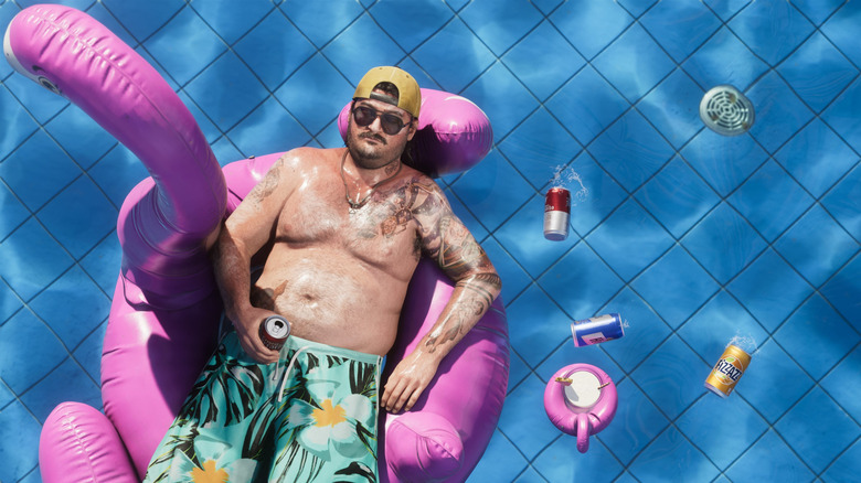 Cal from GTA VI floats on an inflatable pink flamingo in a clear blue pool, beer cans in the water.