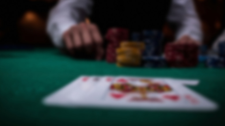Blurred, out-of-focus photo of playing cards face-up on a green felt table, with stacks of chips and a player's arm visible in the background.