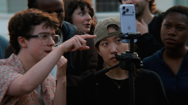 A series of people stood in front of an Apple iPhone in a tripod while taking a video or photo.