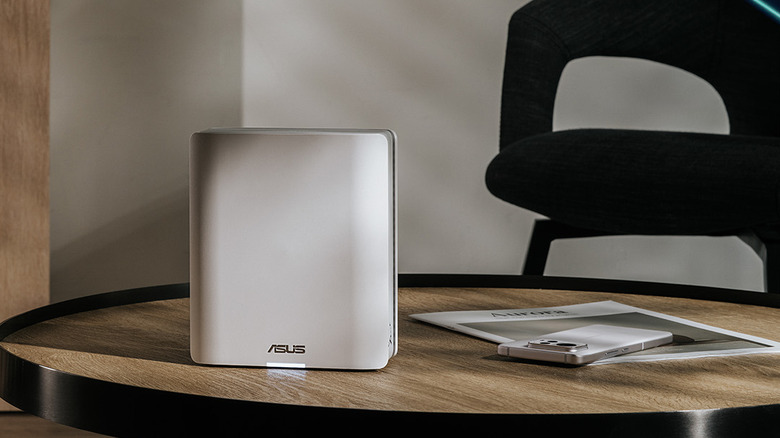 A white ASUS router sits on a table beside a white smartphone and a black chair.