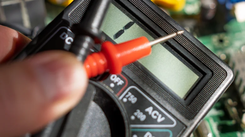 A close-up of a black digital multimeter with a red test probe held beside its display.