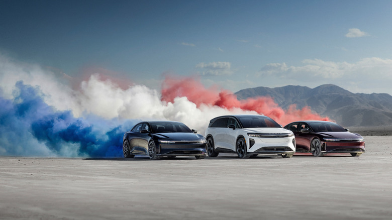 Blue, white, and red Lucid EVs drive together through a desert, churning up a cloud of sand and dust that reflects their respective colors.