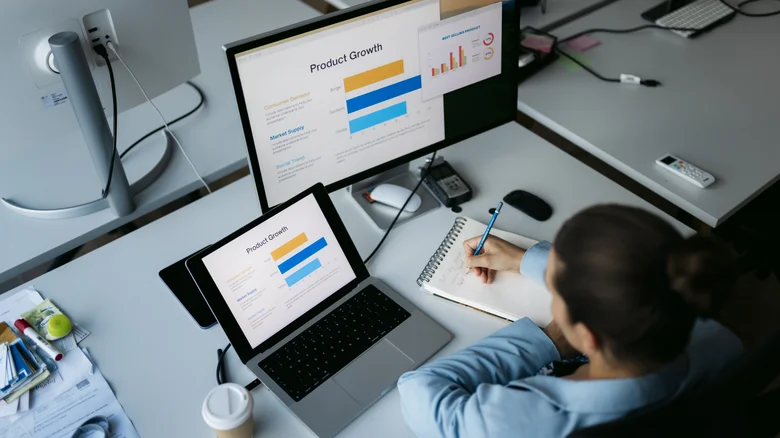 A person works on a laptop connected to a larger external monitor while taking notes at a desk.