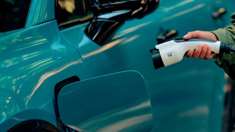 A person moves to plug in a Honda-labeled charger into a bright teal Honda Prologue's charging port.