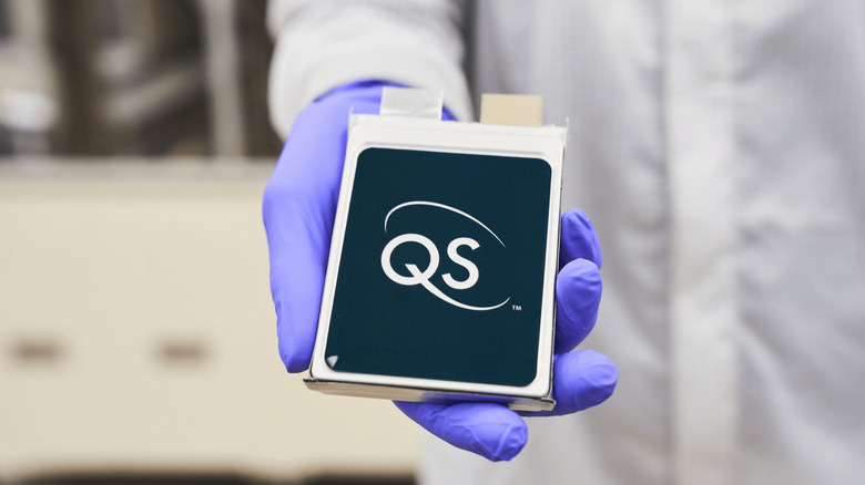 An engineer in a white lab coat and purple gloves shows off a QuantumScape's 24 layer prototype cell, a dark blue and silver battery cell with the company's signature QS symbol on the front.