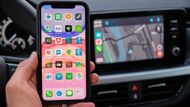 An iPhone in a car, connected by USB cable, with the CarPlay experience shown blurred behind it.