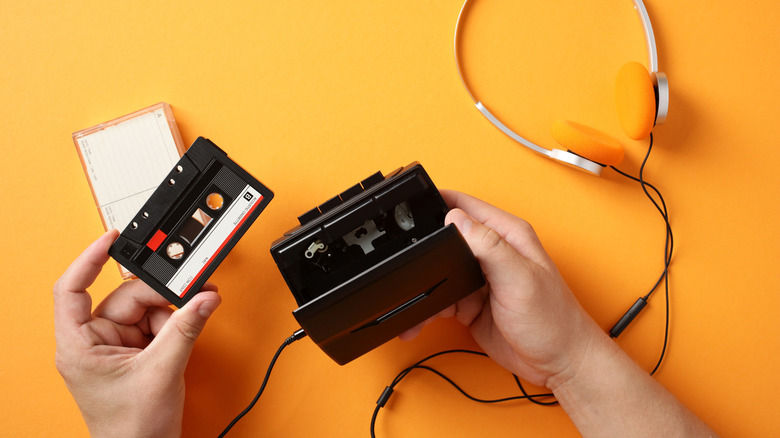A hand holding a cassette tape with an open cassette player on an orange background.