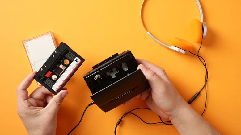 A hand holding a cassette tape with an open cassette player on an orange background.