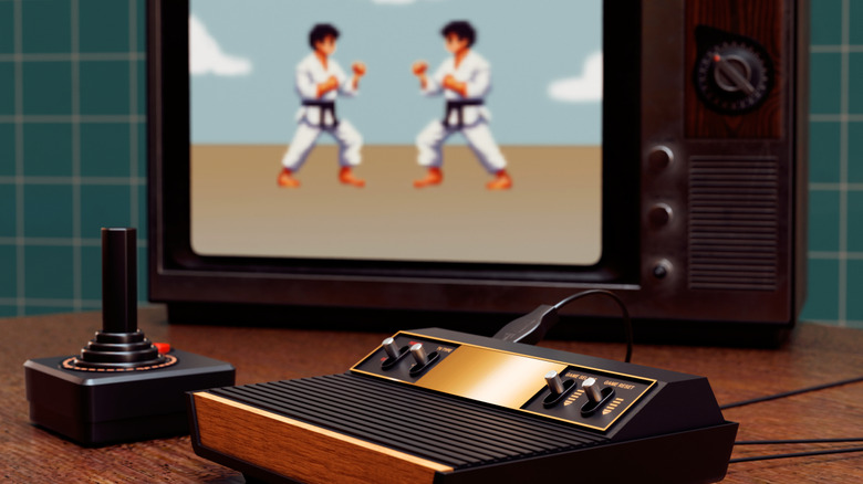 An Atari 2600 video game console and controller with a karate game on an old TV in the background.